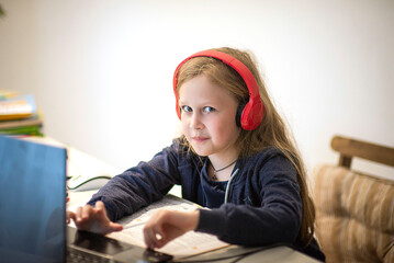 Smiling little European girl with headphones watching video lesson on computer in kitchen, happy little kid with headphones has online web class using laptop at home, home learning concept, selective