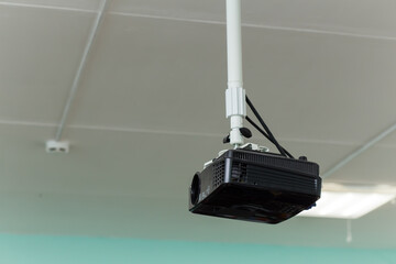 The black video projector is suspended from a long bracket under the ceiling. Against the background of light ceiling panels. Signal and power cables are connected at the top.