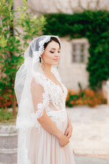 A gentle bride in a lace veil stands near a white house entwined with a vine in the old town of Perast 