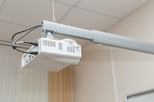 The white video projector is suspended from an adjustable ceiling bracket. Against the background of light ceiling panels. Signal and power cables are connected from the rear.