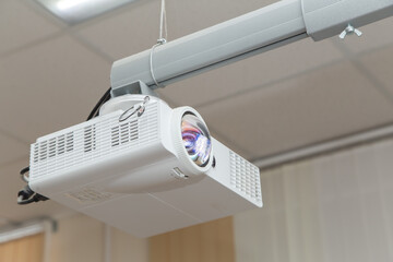 A white video projector hangs from the ceiling with an adjustable arm. Against the background of light ceiling panels. Round objective lens with abstract pattern.