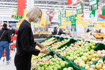 Masked woman in the vegetable section of the supermarket. Vitamins and healthy eating during the coronavirus pandemic.