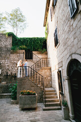 Fototapeta na wymiar The bride and groom stand on a beautiful staircase with a wrought iron railing in the old town of Perast and look at each other 