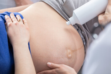 A nurse squeezes gel onto a pregnant patient's abdomen and prepares for an ultrasound. Gynecology office.