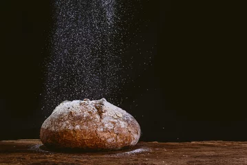 Fototapeten rustic and golden round loaf of fresh whole grain bread on dark black background on top of wooden kitchen table with cascade of falling flour © davide bonaldo
