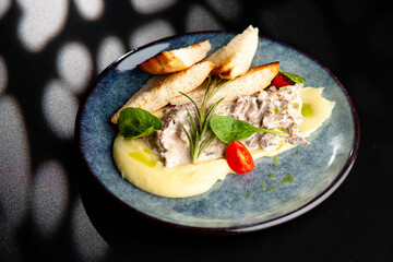 Meat in a creamy sauce with mashed potatoes and bread toast, painted with spinach leaves and cherry tomatoes, on a gray plate on a black background.