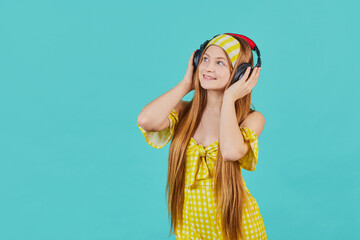 Studio portait of Funny young blonde woman in yellow dress isolated on blue background. Girl Listen music with wireless headphones using music streaming service.