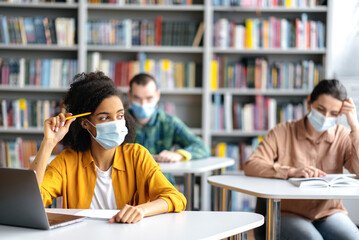 Multiracial students in protective medical masks sit at tables at a safe distance in the library during a lecture, a smart african american woman looks away and thinks about lecture or weekend