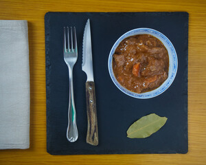 Fricando, Traditional Catalan tapa, stew of beef in a bowl decorated with metal cutlery, a napkin and a bay leaf on a black slate board. Top view