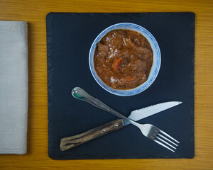 Fricando, Traditional Catalan tapa, stew of beef in a bowl decorated with metal cutlery, a napkin on a black slate board. Top view