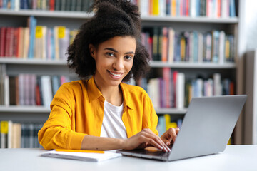 Pretty young african american teenage girl, trendy dressed, female student sitting in the campus library at the laptop, preparing to the exam, searching information, looking at the camera, smiling