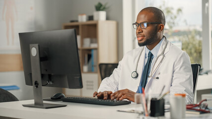 Fototapeta na wymiar Experienced African American Male Doctor Wearing White Coat Working on Personal Computer at His Office. Medical Health Care Professional Working with Test Results, Patient Treatment Planning.