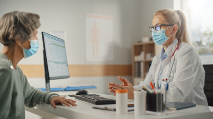 Female Family Doctor is Talking with Senior Old Patient During Consultation in a Health Clinic. Both Wear Face Masks. Physician in Lab Coat Sitting Behind a Computer Desk in Hospital Office.