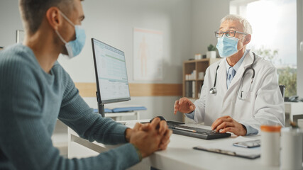 Middle Aged Family Doctor is Talking with Young Male Patient During Consultation in a Health Clinic. Both Wear Face Masks. Physician in Lab Coat Sitting Behind a Computer Desk in Hospital Office. - Powered by Adobe