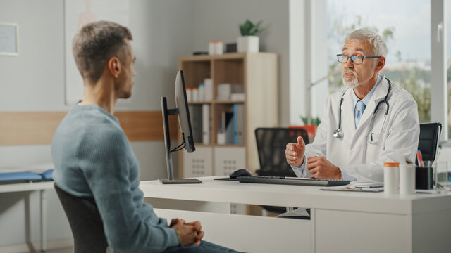 Middle Aged Family Doctor Is Talking With Young Male Patient During Consultation In A Health Clinic. Senior Physician In Lab Coat Sitting Behind A Computer Desk In Hospital Office.