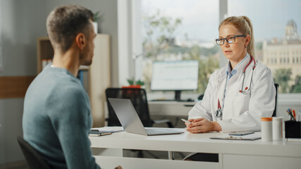 Female Family Doctor is Talking with Healthy Young Male Patient During Consultation in a Health Clinic. Experienced Physician in Lab Coat Sitting Behind a Computer Desk in Hospital Office.