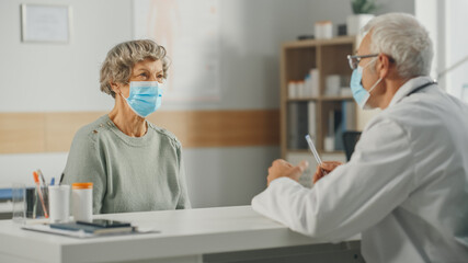 Obraz premium Middle Aged Family Doctor is Talking with Senior Female Patient During Consultation in a Health Clinic. Both are Wearing Face Masks. Physician in Lab Coat Sitting Behind a Desk in Hospital Office.