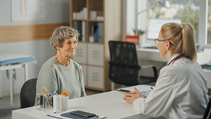Female Family Doctor is Sitting Behind a Desk and Speaking to a Senior Woman During Consultation in a Health Clinic. Physician in Lab Coat Prescribing Drugs to Elderly Patient in Hospital Office.