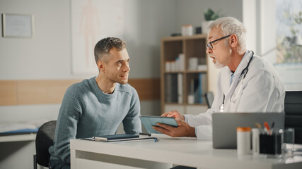 Experienced Middle Aged Family Doctor Showing Analysis Results on Tablet Computer to Male Patient During Consultation in a Health Clinic. Physician Sitting Behind a Desk in Hospital Office. - Powered by Adobe
