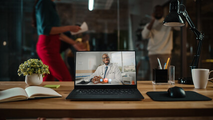 Shot of a Laptop Computer with Portrait of a Friendly Male Medical Consultant Having Online Video Call. Computer on a Desk in a Busy Creative Office Environment. Authentic Hipster Agency Vibe. 