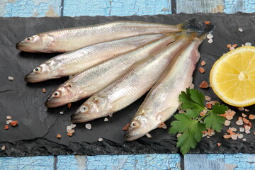 Fresh smelt fish on a black stone plate and wooden background with old blue color, Smelt fishes (European smelt) with lemon and green parsley, Group of fresh smelt fish 