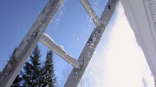 POV under a ladder leaning on the edge of a roof, shooting shovelfuls of snow falling as someone on the roof throws them down. Sunny winter day