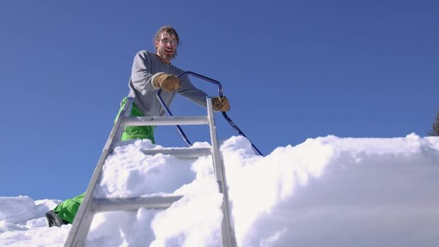 Shooting from below of a young man on the roof of a house, pushing heaps of snow over the edge with a shovel, towards camera. Sunny winter day.