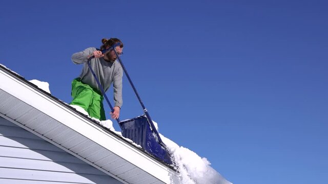 Wide angle view of an athletic young man on the roof of a house in a bright sunny winter day shoveling down heaps of fresh snow near the edge.