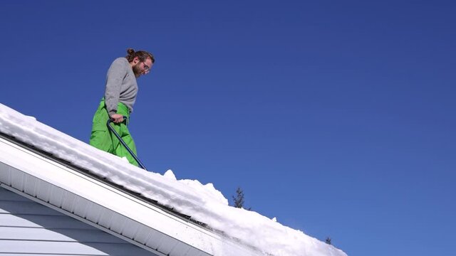 Wide angle view of a strong young man on the roof of a house in a bright sunny winter day shoveling down heaps of fresh snow with a hand tool.