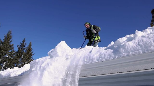 Shooting from ground level of a young man perched on the roof of a house removing fresh snow with a shovel and throwing it downwards.