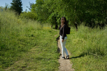 beautiful long-haired woman in black shirt, white pants and brown bag walking through nature with trees in the background and blue sky. teenager walking in the park watching the camera