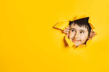 Little caucasian boy looks in the gap in a colored yellow paper wall. The child looks at the empty space on the side. A place for text and promotional information.