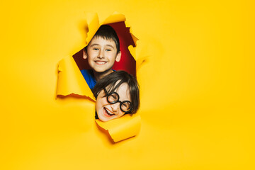 Two funny and laughing kids look into a torn hole in a colored paper wall. Advertising banner for...