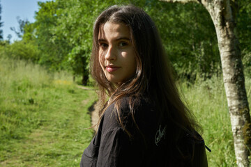 beautiful long-haired woman in black shirt, white pants and brown bag walking through nature with trees in the background and blue sky. teenager walking in the park watching the camera