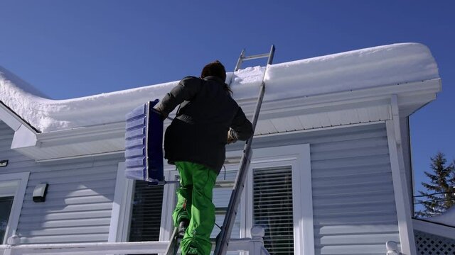 Long haired young man in fur hat carefully climbing with a tool an extensible ladder to reach the roof of a house for a snow removal operation.
