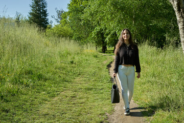 beautiful long-haired woman in black shirt, white pants and brown bag walking through nature with trees in the background and blue sky. teenager walking in the park watching the camera