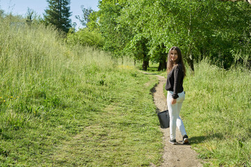 beautiful long-haired woman in black shirt, white pants and brown bag walking through nature with trees in the background and blue sky. teenager walking in the park watching the camera