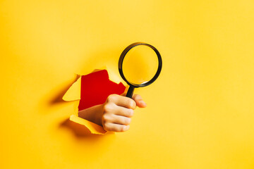 A human hand holds a magnifying glass through a torn hole in a paper yellow wall. Search...