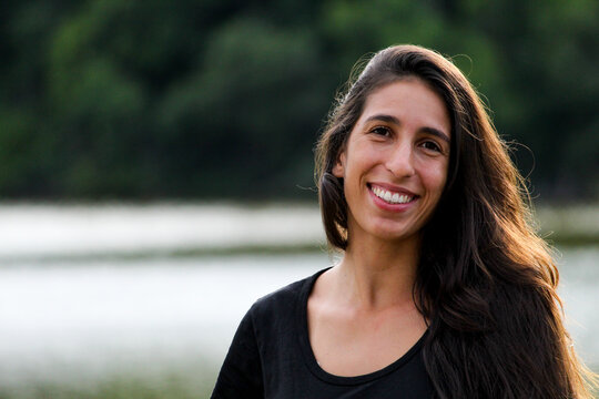 MULHER JOVEM, SORRINDO, LAGOA DESFOCADA NO FUNDO.