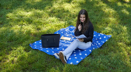 beautiful long-haired woman writing in a brown notebook in nature sitting on the grass with a blanket and her bag with trees in the background. teenage girl drawing in a notebook in the park smiling