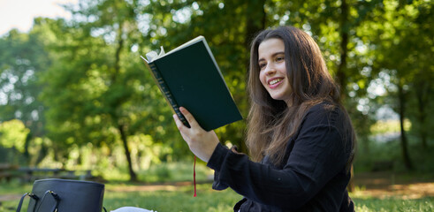 beautiful long-haired woman reading a green book in nature sitting on the grass with a blanket and her bag with trees in the background. teenager looking at a literary work in the park smiling