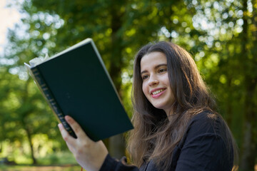 beautiful long-haired woman reading a green book in nature sitting on the grass with a blanket and her bag with trees in the background. teenager looking at a literary work in the park smiling