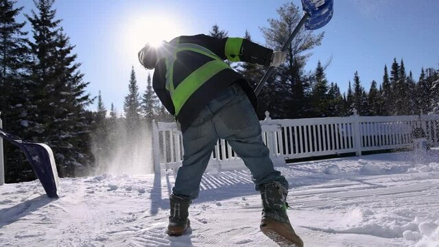 Snow removal in a sunny winter day. Strong young man in fluo visibility jacket shoveling fresh snow from a first floor veranda with a blue shovel.