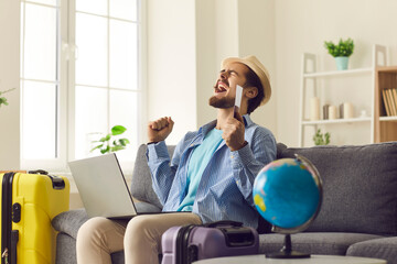 Online travel agency concept. Young happy excited male traveller in hat screaming doing winner gesture clenching fists say yes holding passport with flight tickets sitting front of laptop on home sofa