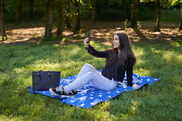 young woman with long hair in a black shirt and white pants sitting on a blanket of stars on the grass in a park with her bag using her phone to take pictures and video conferencing. influencer nature