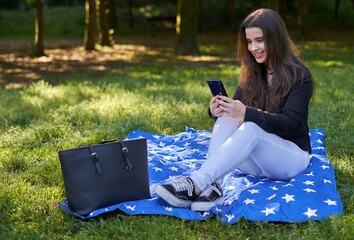young woman with long hair in a black shirt and white pants sitting on a blanket of stars on the grass in a park with her bag using her phone to take pictures and video conferencing. influencer nature