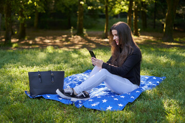 young woman with long hair in a black shirt and white pants sitting on a blanket of stars on the grass in a park with her bag using her phone to take pictures and video conferencing. influencer nature