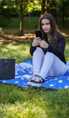 young woman with long hair in a black shirt and white pants sitting on a blanket of stars on the grass in a park with her bag using her phone to take pictures and video conferencing. influencer nature