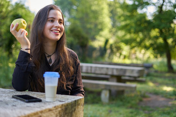 beautiful young woman in a black shirt sitting on a stone table eating an apple in nature with her mobile, a coffee and her bag with trees in the background. vegan teen taking a break in the park