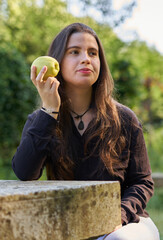 beautiful young woman in a black shirt sitting on a stone table eating an apple in nature with her mobile, a coffee and her bag with trees in the background. vegan teen taking a break in the park